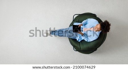 People And Modern Technology. Above top view of young smiling black lady holding tablet sitting on bean bag in living room, watching video. Cheerful lady surfing internet, free copy space, panorama Royalty-Free Stock Photo #2100074629