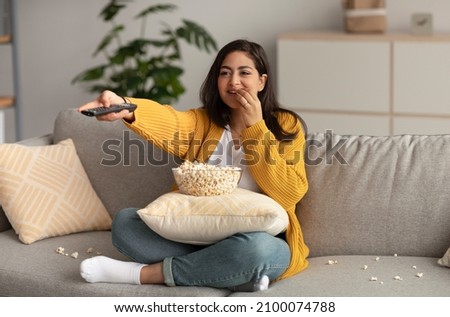 Young middle eastern lady spending her free time at home, watching TV and eating popcorn on sofa, free space. Excited arab lady with remote control enjoying interesting movie Royalty-Free Stock Photo #2100074788