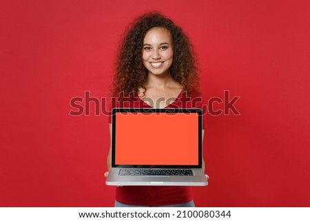Smiling young african american woman girl in casual t-shirt posing isolated on red background studio. People lifestyle concept. Mock up copy space. Hold laptop pc computer with blank empty screen
