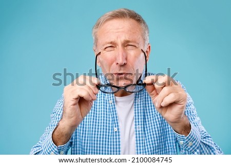 Poor Eyesight. Mature Man Can't See, Squinting Eyes Putting On Glasses Having Problems With Vision, Looking At Camera At Blue Studio. Ophtalmic Issue, Bad Sight In Older Age, Macular Degeneration Royalty-Free Stock Photo #2100084745