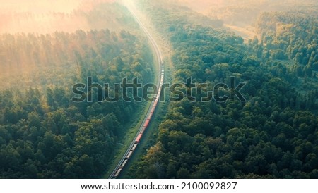 Cargo Train in summer morning forest at fog sunrise. Aerial view of moving freight train in forest. Morning mist landscape with train, railroad, foggy trees. Top aerial drone view near railway. Royalty-Free Stock Photo #2100092827