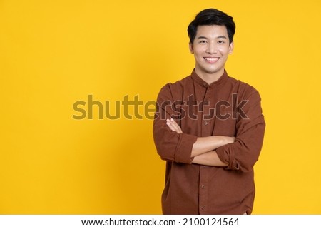 Smiling young Asian man with arms crossed in yellow studio isolated background Royalty-Free Stock Photo #2100124564