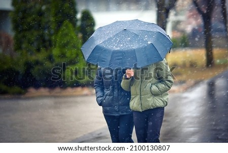 Two mid aged women walking under umbrella during snowfall in city street, Selective focus. Female friends with one umbrella in cold  snowy winter season walking on street. Couple under umbrella