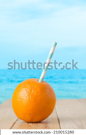 A whole orange, pierced with a striped straw to represent a fresh fruit drink.  Set on a planked wooden table with soft focus sea and sky background.