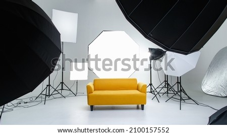 Studio shot fashion backstage photographing shooting set with yellow cozy sofa couch and photography equipment softbox flash strobe spotlight tripods reflector umbrella on white backdrop background. Royalty-Free Stock Photo #2100157552
