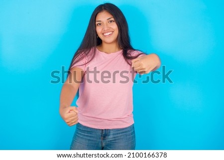 Portrait of charming Young hispanic girl wearing pink T-shirt over blue background, smiling broadly while holding hands over her head.  Confidence and relax concept.