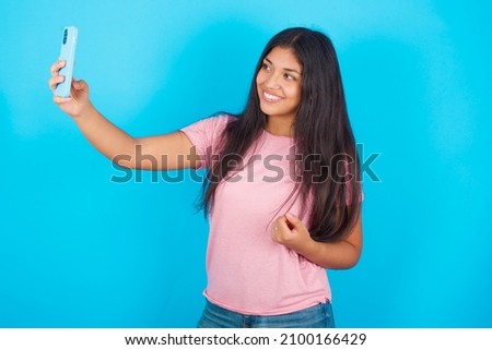 Portrait of a Young hispanic girl wearing pink T-shirt over blue background  taking a selfie to send it to friends and followers or post it on his social media.