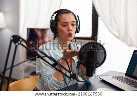 Woman influencer blowing kiss on microphone on live podcast. Content creator with headphones recording online broadcast in home studio. Internet presenter hosting radio livestream.