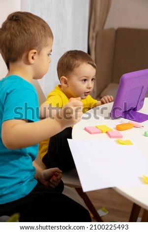 Happy kids with yellow and blue shirts doing arts and crafts together at their desk and watching cartoon on digital tablet. Children have fun. Boys play at home.