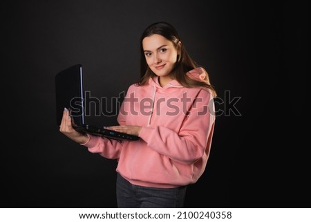 A student or a freelancer with a laptop in her hands. A young charming woman in a pink hoodie works remotely. Brunette holds an open laptop, photo on black