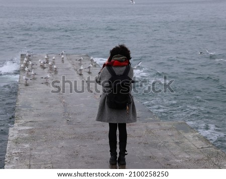 curly girl with a backpack in a coat takes pictures of seagulls by the sea