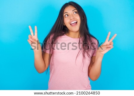 Isolated shot of cheerful Young hispanic girl wearing pink T-shirt over blue background  makes peace or victory sign with both hands, feels cool.