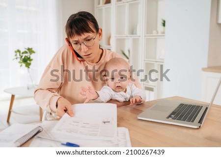 Busy single mother taking phone call and trying to solve family promlems while taking care of her little baby. Multitasking stressed mom talking on cellpphone, sitting at desk, holding her toddler Royalty-Free Stock Photo #2100267439