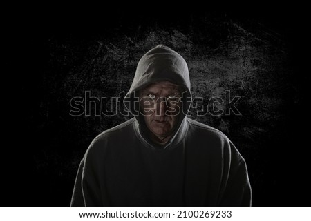 An evil looking man with piercing eyes and a hoodie stands intimidatingly against an ally wall stalking his next victim.     Royalty-Free Stock Photo #2100269233