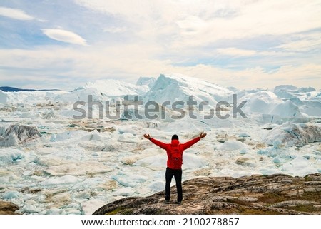 Climate Change and Global Warming Optimism and Positivity Concept. Man on Greenland arms raised in awe of iceberg nature. Melting of glaciers and Greenland ice sheet is a cause of sea levels rise. Royalty-Free Stock Photo #2100278857