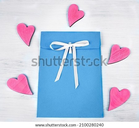 Blue envelope with a white bow lies on a white wooden table. There are a lot of bright pink hearts around