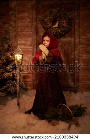 Once upon a Christmas. A cute little girl in medieval clothes stands by a wooden house and burning lantern on a cold night with a basket of fir branches brought from the forest. Full length shot.