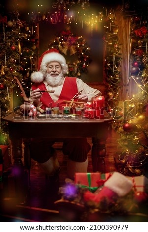 Portrait of jolly Santa Claus at his work in the workshop making and painting toys for gifts to children. Beautiful room festively decorated in honor of Christmas. Christmas and New Year concept. 