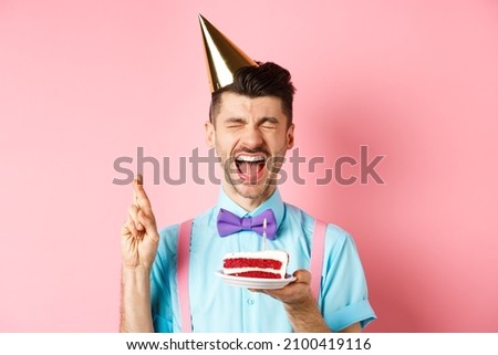 Holidays and celebration concept. Excited guy celebrating birthday and making wish, cross fingers for good luck while holding bday cake with candle, pink background