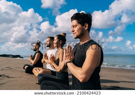 Young male and female in lotus pose holding hands in namaste enjoying spiritual meditation at coastline beach, group of friends have togetherness morning yoga practice at seashore in Indonesia Royalty-Free Stock Photo #2100424423