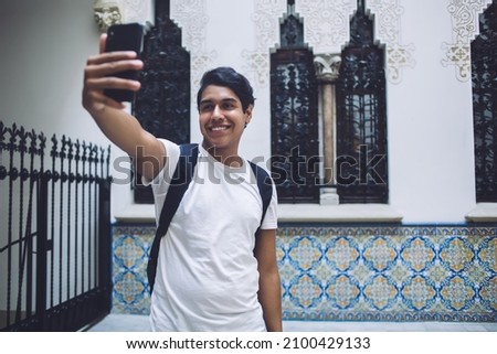Toothy male tourist using front camera on mobile device for clicking funny photo content during getaway travelling, cheerful hipster guy shooting web vlog about international trip on vacations