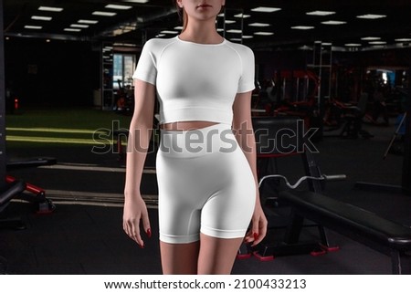 Stylish crop top mockup, t-shirt, high workout shorts, blank sportswear on sportswoman posing against the backdrop of the gym. Compression suit template for yoga, run, fitness, place for design, brand Royalty-Free Stock Photo #2100433213