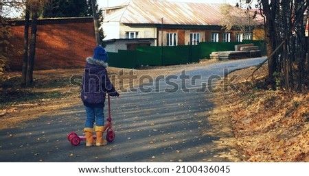 A girl in a blue hat, riding a pink scooter in the fall. Asphalted road, sports. Yellow rubber boots. Autumn, spring, seasons.