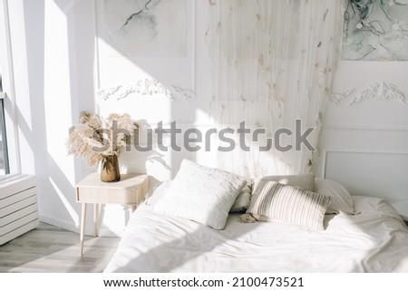 White cozy bedroom  where the sun shines from the window. White bedding, dried flowers in a vase on a bedside table for decoration. Royalty-Free Stock Photo #2100473521