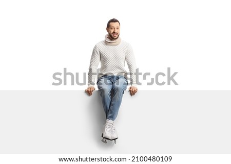 Casual young man wearing ice skates and sitting on a blank panel isolated on white background