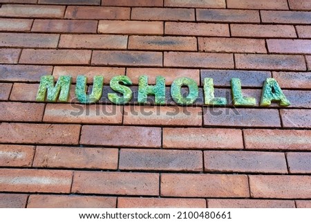 the wall of a place  room of worship for Muslims with the words "MUSHOLLA", - as a marker it is a place of worship for Muslims.the writing is made of stone which gives a natural impression in harmony