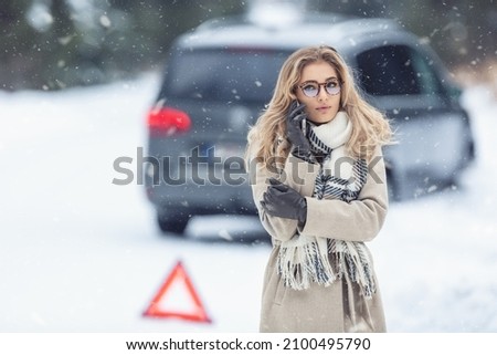 Inexprienced female driver who crashed the car off the slippery winter road calls towing service with emergency triangle next to her outside the car.