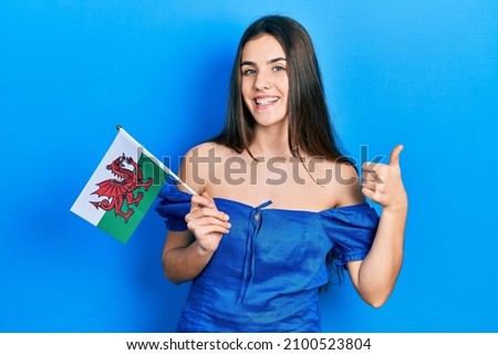 Young brunette teenager holding wales flag smiling happy and positive, thumb up doing excellent and approval sign 