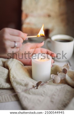 Female hands with lit match lighting burning candle on windowsill for calm and coziness at home, woman trying to create cozy warm and intimate atmosphere in cold season. Hygge lifestyle concept Royalty-Free Stock Photo #2100544945