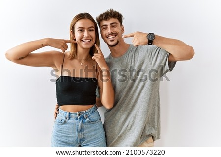 Young beautiful couple standing together over isolated background smiling cheerful showing and pointing with fingers teeth and mouth. dental health concept.  Royalty-Free Stock Photo #2100573220