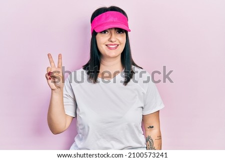 Young hispanic woman wearing sportswear and sun visor cap smiling with happy face winking at the camera doing victory sign. number two.  Royalty-Free Stock Photo #2100573241