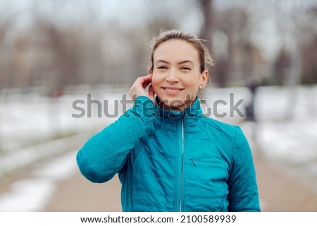 Portrait of sportswoman in warm outfit standing in public park on snowy weather and putting earphones. Winter sport, music, recreation, technology