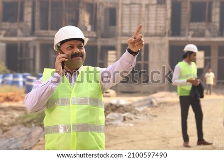 Hard working male construction engineer standing on a construction site. The engineer in wearing helmet and uniform showcasing work in progress. Talking on phone and pointing upwards.