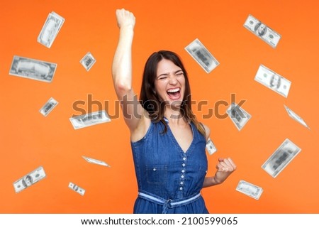 Money rain, winner and rich. Hurray. satisfied happy young woman rising hands up with toothy smile on face, pleased, amazed with money falling. Indoor studio shot isolated on orange background Royalty-Free Stock Photo #2100599065
