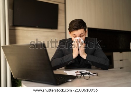 Sick young man at work got allergic to flu, sneezing and fasting, wiping a runny nose with a paper napkin, allergic guy caught a cold at work, respiratory illness sitting in home office, sick leave Royalty-Free Stock Photo #2100610936