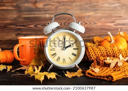 Alarm clock and autumn leaves with cup of tea and clothes on wooden background. Daylight saving time end Royalty-Free Stock Photo #2100637987