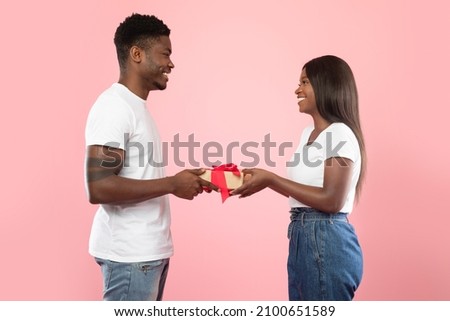 Happy black couple giving gifts to each other, pink studio background, free space. Loving handsome man giving present to his excited wife or girlfriend. Valentines day celebration, profile side view