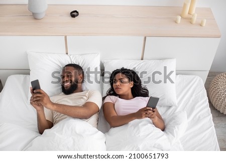 Disgruntled millennial african american wife looks at husband smartphone on bed in bedroom interior, top view. Gadget addiction, online games and social networks, treason and relationship problems