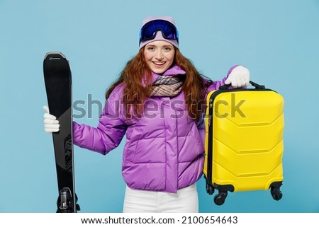 Traveler tourist woman wear winter ski windbreaker jacket goggles hold valise suitacase isolated on plain blue background studio. Passenger travel abroad on weekends getaway Air flight journey concept