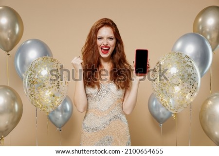 Young redhead woman in evening dress balloons hold mobile cell phone with blank screen workspace area do winner gesture isolated on plain pastel beige background. Celebrating birthday party concept