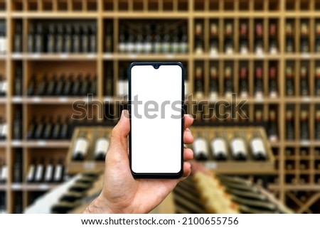 Wine shop with bottles background. Hand with blank smartphone screen one wine backdrop. Buying and ordering alcoholic beverages online