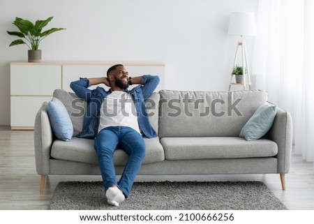 Cheerful attractive millennial african american male with beard sitting on couch and look at empty space in minimalist living room interior. Rest at home, break. Man enjoys weekend and free time Royalty-Free Stock Photo #2100666256