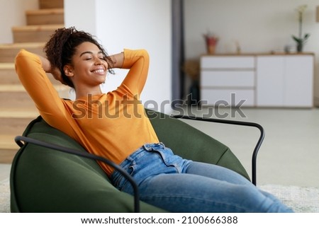 Rest Concept. Happy black lady sitting on comfortable bean bag at home in living room. Cheerful casual woman with closed eyes relaxing on sofa, leaning back, enjoy weekend free time or break from work Royalty-Free Stock Photo #2100666388