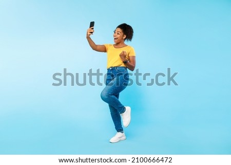 Full length portrait of emotional young black woman looking at smartphone screen, making YES gesture, jumping in excitement, winning online lottery, achieving success on blue studio background Royalty-Free Stock Photo #2100666472