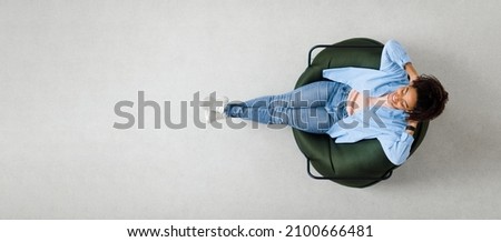 Rest Concept. Above top view of happy black lady sitting on bean bag at home in living room. Cheerful casual woman relaxing on sofa leaning back, enjoying free time, banner panorama, free space mockup Royalty-Free Stock Photo #2100666481