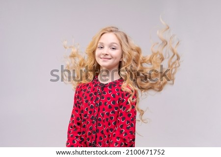 A beautiful cheerful girl in red dress with flying long wavy hair on light gray background laughs with joy. Hair care lightness and beauty of children's hair. Caucasian young girl